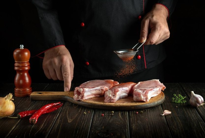 Chef seasoning raw pork chops with paprika using a fine strainer.