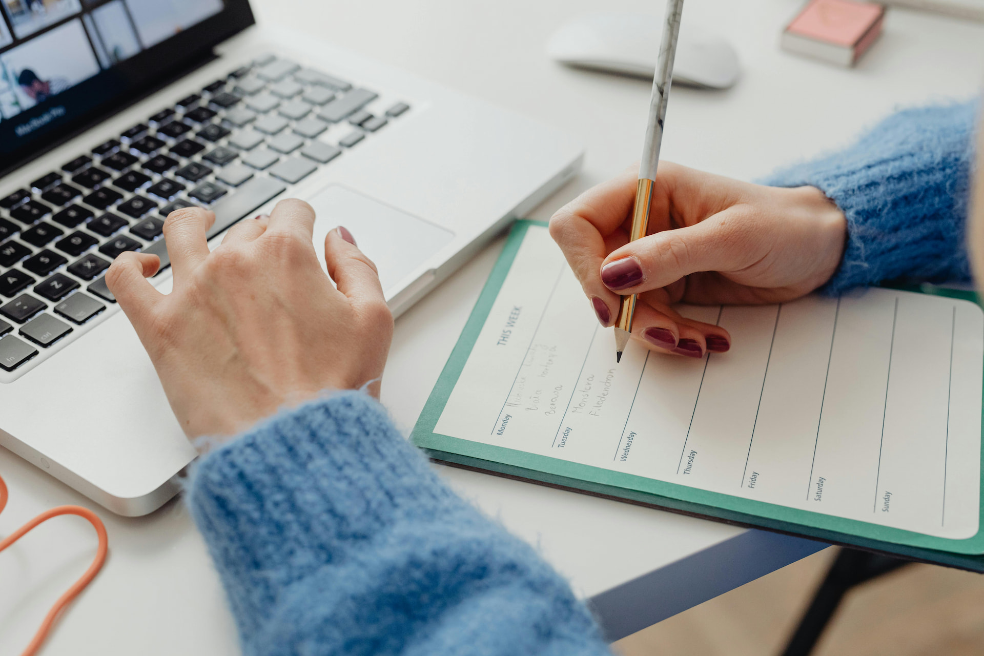 A woman writing notes on a notebook while looking at laptop screen.